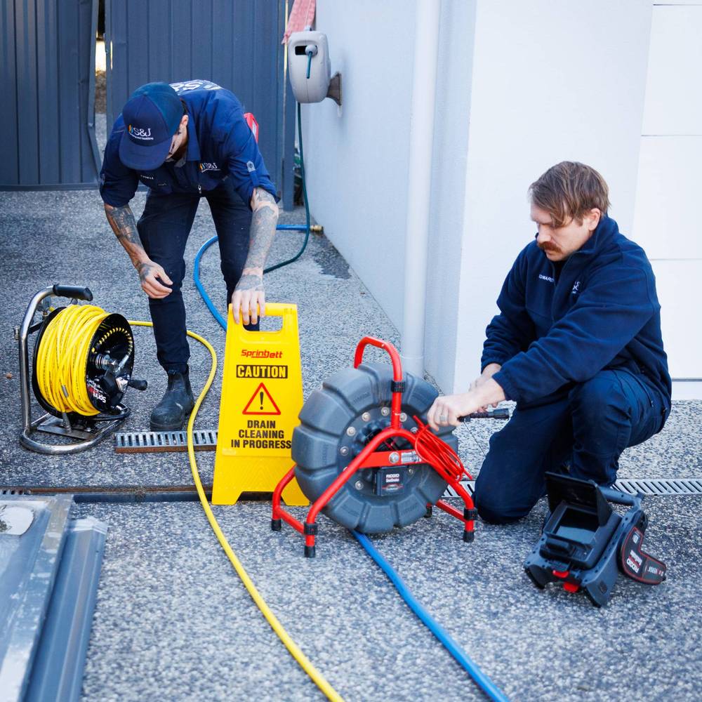 Two S&J plumbers setting up a CCTV drain camera and a high-pressure jetter beside a drain, with a drain cleaning in progress sign
